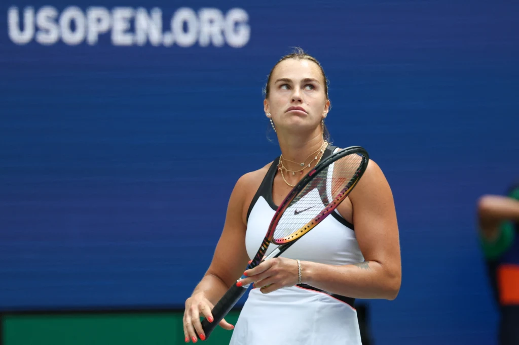 Aryna Sabalenka, US Open 2025 Tenisistka w sportowym stroju trzymająca rakietę na tle niebieskiego banneru z napisem USOPEN.ORG, skoncentrowana, otoczona sportową atmosferą turnieju.