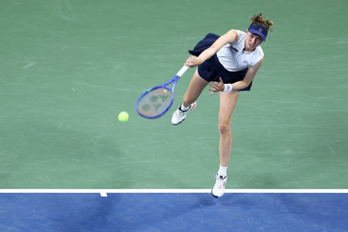 Tenisistka w sportowym stroju wykonuje dynamiczny serwis na korcie o zielonej nawierzchni, skupienie na twarzy, piłka tuż przed rakietą, akcja uchwycona w ruchu.