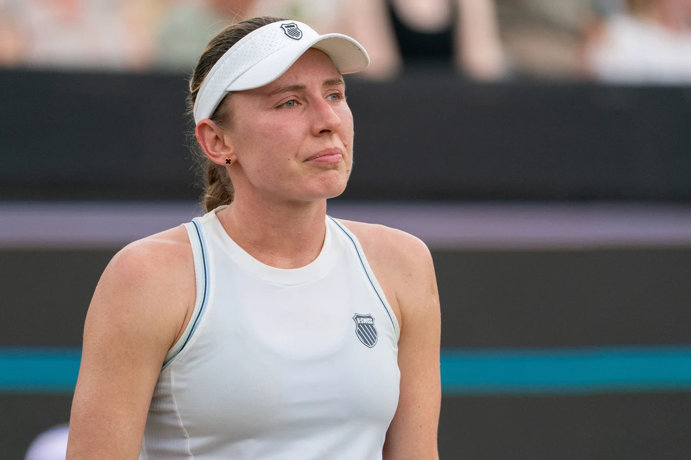 Kobieta w sportowym stroju i białej czapce z daszkiem na korcie tenisowym, wyraz twarzy skupiony i lekko zamyślony, w tle widoczne rozmazane postaci.