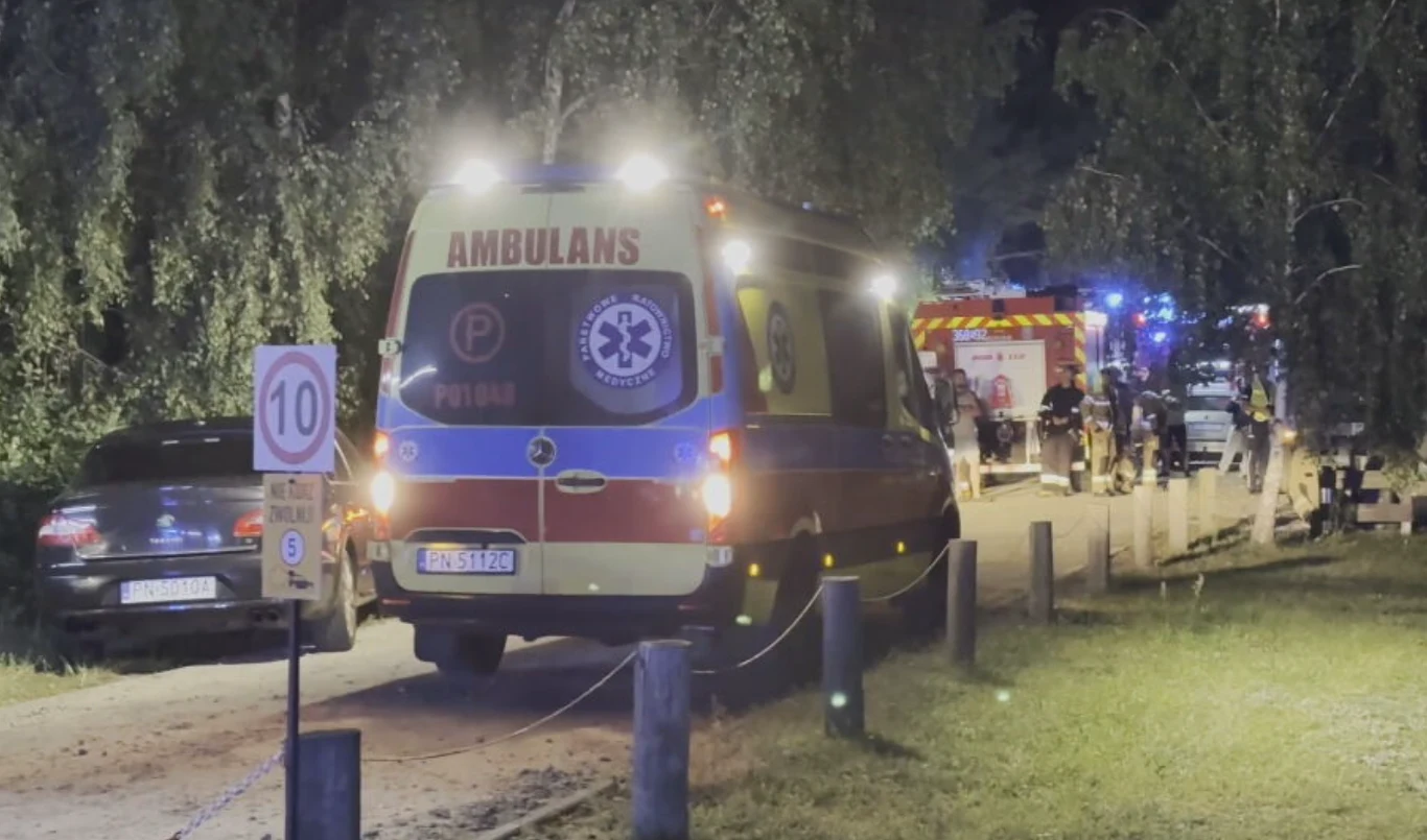 Tragedia w Helenowie Pierwszym Stojący ambulans i w oddali samochód straży pożarnej z włączonymi światłami na leśnej drodze, widoczne inne pojazdy ratunkowe, otoczenie nocne, delikatnie oświetlone przez lampy pojazdów.