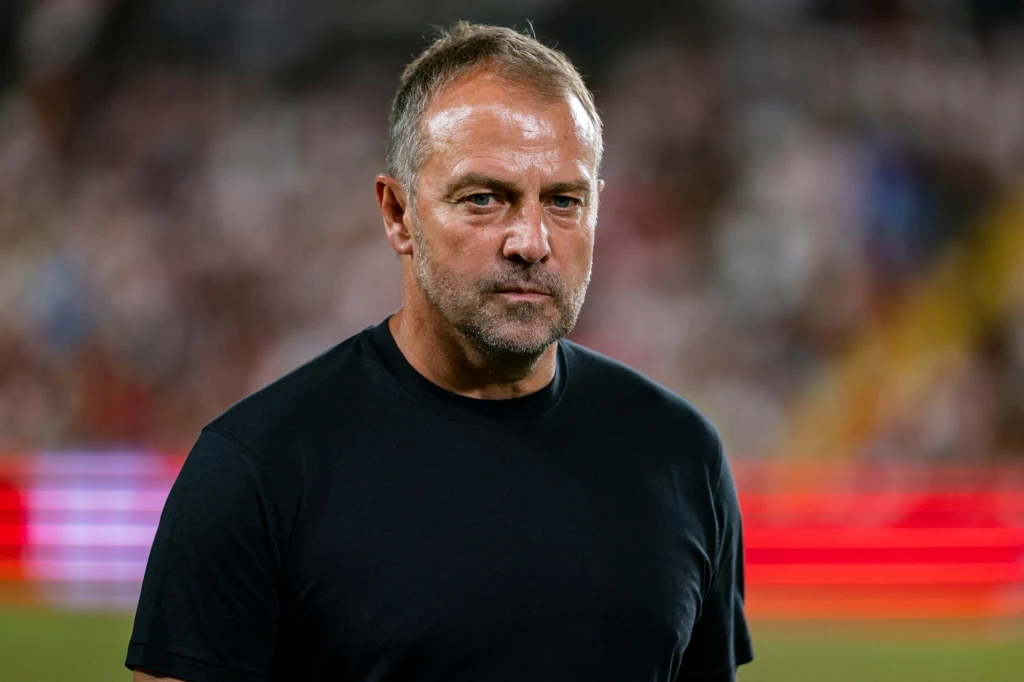 Hansi Flick Mężczyzna w średnim wieku w ciemnej koszulce stoi na tle rozmytego stadionu z rozświetlonymi czerwonymi panelami świetlnymi w tle, spogląda poważnie w stronę kamery.