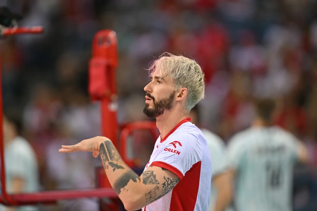 Tomasz Fornal Zawodnik drużyny siatkarskiej w biało-czerwonym stroju, z widocznymi tatuażami na ramieniu, wykonuje gest ręką podczas meczu, w tle rozmyci kibice i inni zawodnicy.
