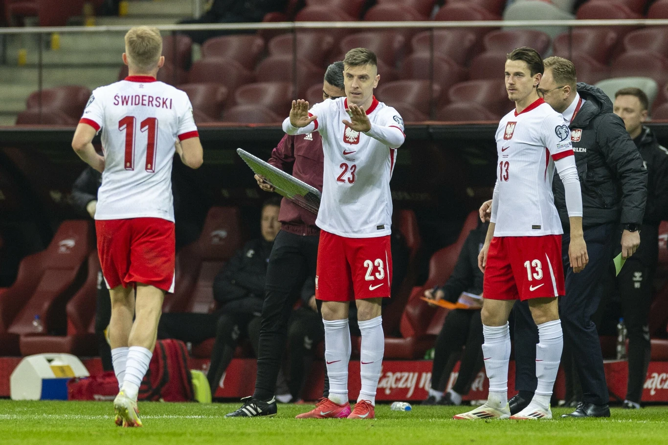 Piłkarz o numerze 23 przygotowuje się do wejścia na boisko podczas meczu, obok niego stoi drugi zawodnik w stroju reprezentacji Polski, a zawodnik z numerem 11 opuszcza boisko, całość rozgrywa się na stadionie.