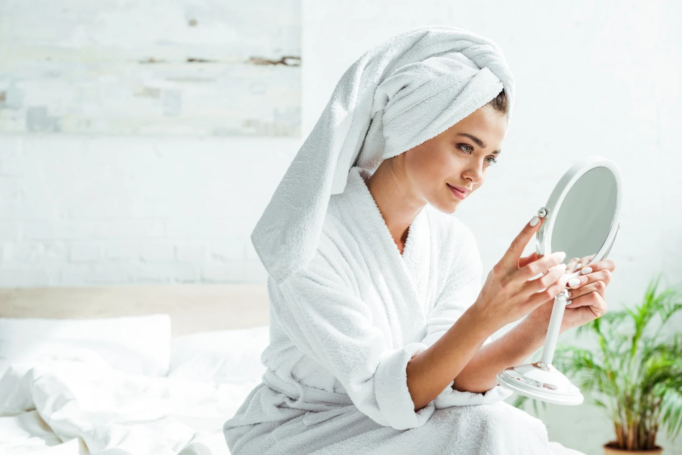 Młoda kobieta w białym szlafroku siedzi na łóżku z ręcznikiem owiniętym na głowie, trzyma w dłoni okrągłe lusterko i z uwagą przygląda się swojemu odbiciu; w tle jasny, minimalistyczny pokój z zieloną rośliną.