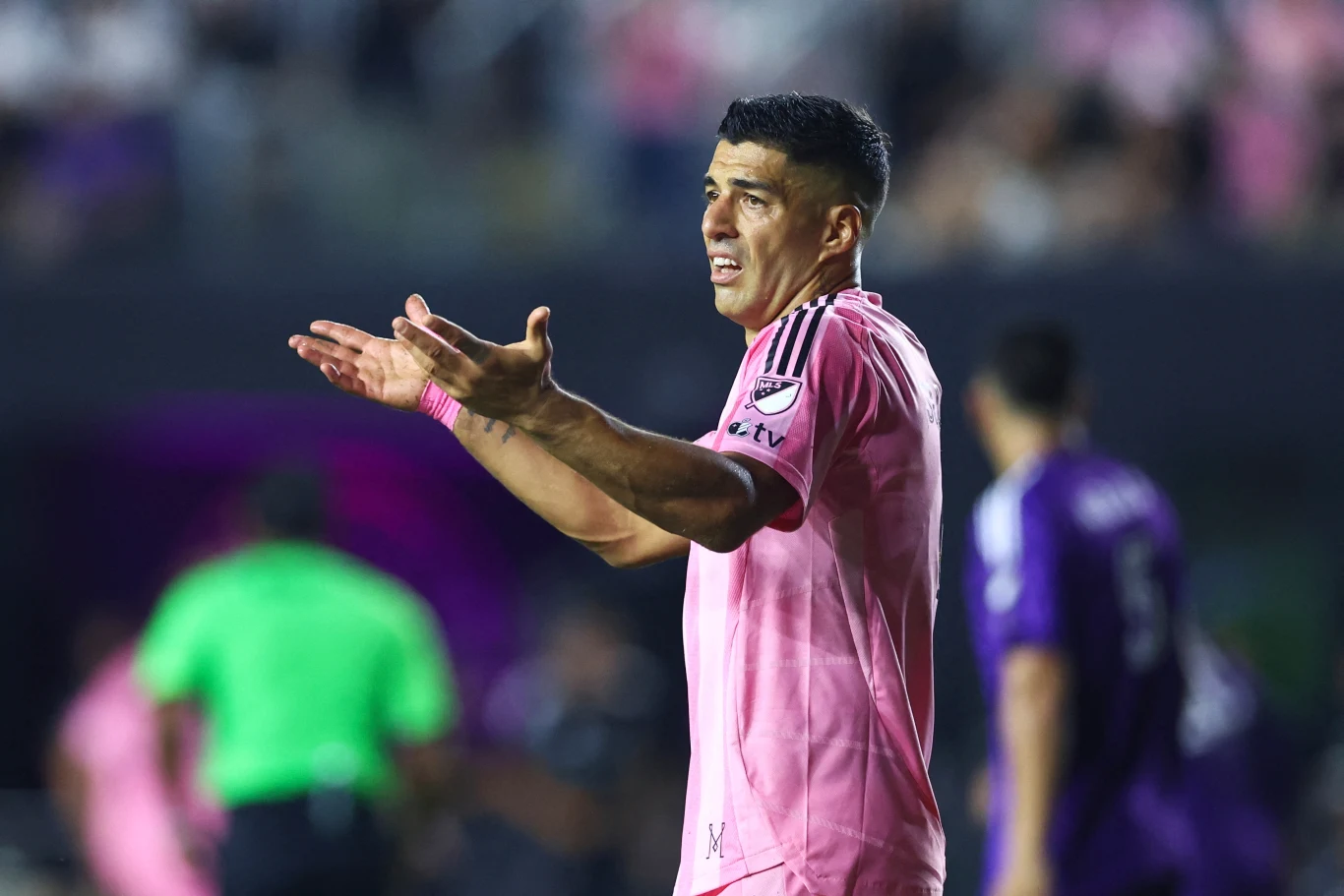 Luis Suarez Piłkarz w różowej koszulce gestykuluje z wyraźną dezaprobatą na boisku podczas meczu, w tle widoczni są inni zawodnicy oraz sędzia.