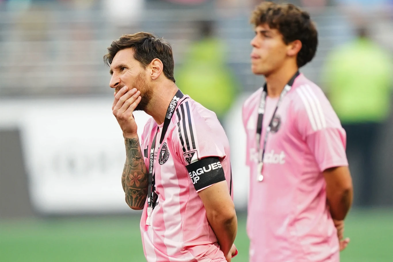 Lionel Messi Dwóch piłkarzy w różowych strojach piłkarskich stoi na murawie po zakończonym meczu, jeden z nich trzyma rękę przy twarzy i wygląda na zamyślonego, na ich szyjach zawieszone są medale.