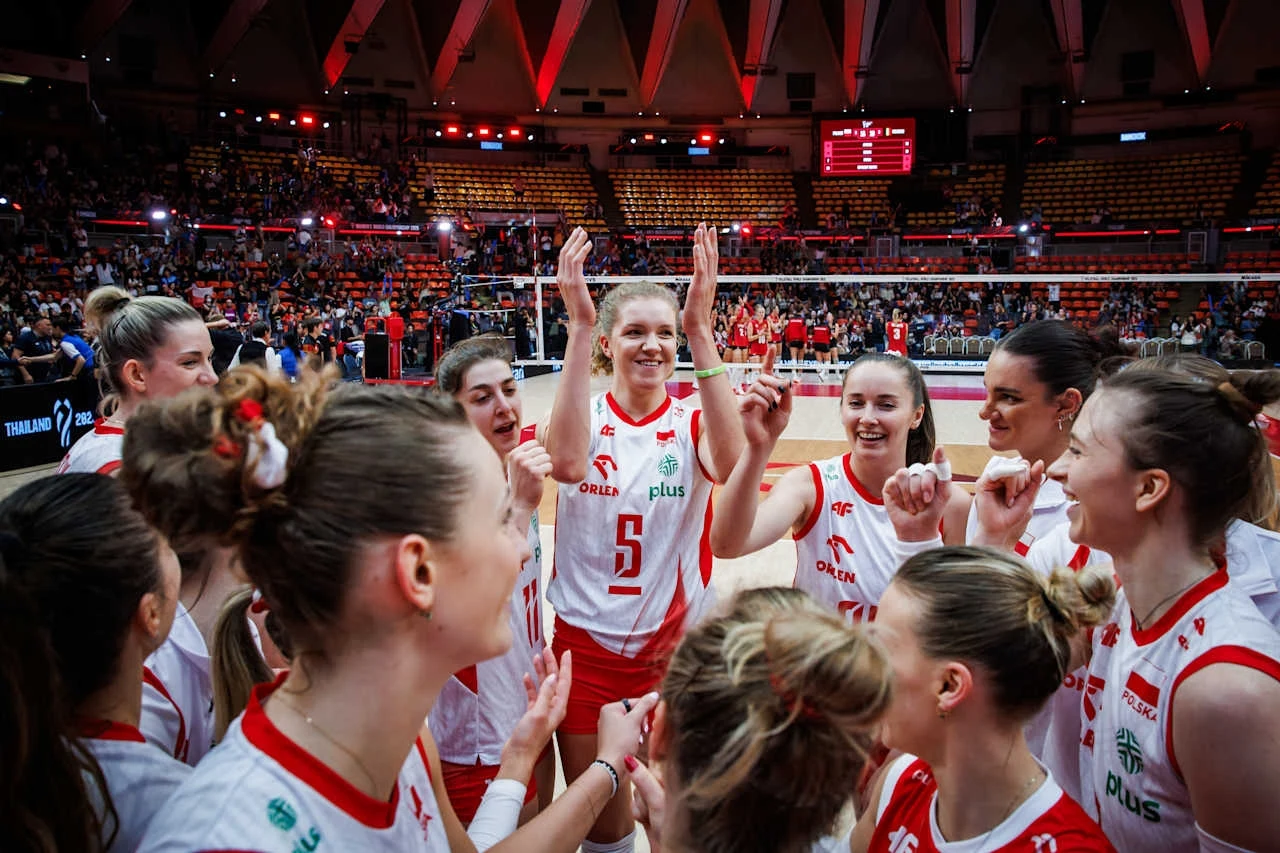 Drużyna siatkarek w białych i czerwonych strojach, skupiona w kole podczas radosnego momentu, na pierwszym planie widoczna zawodniczka z podniesionymi rękami, wystrój hali sportowej wypełnionej widownią i jasnym oświetleniem.