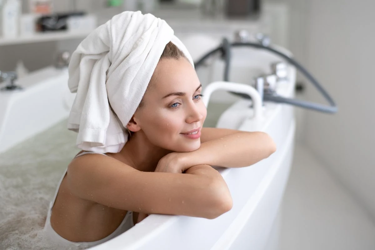 „Po tej kąpieli twoja skóra odzyska miękkość i blask Młoda kobieta z turbanem z ręcznika na głowie relaksuje się w wannie wypełnionej wodą, opierając się o rant wanny i patrząc z uśmiechem w dal, w tle widoczne są elementy łazienki.