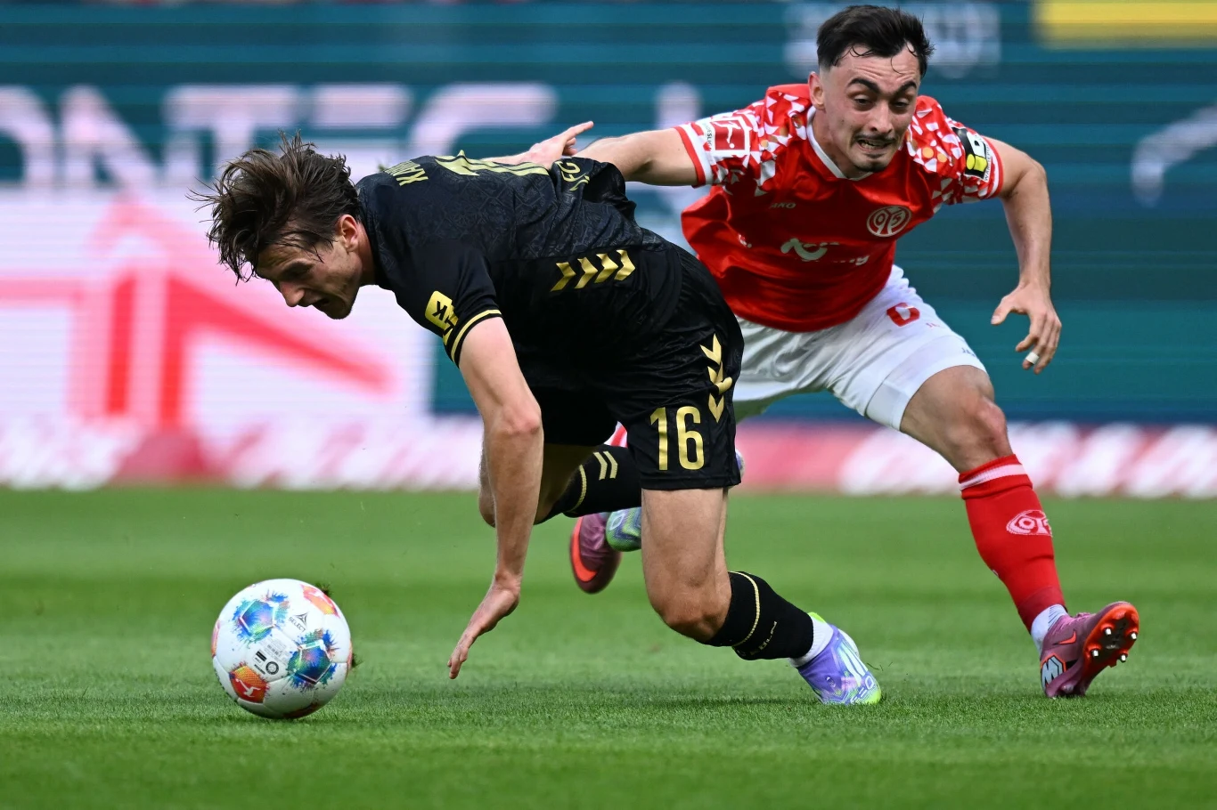 Dwóch piłkarzy podczas zaciętej walki o piłkę na murawie, zawodnik w czarnej koszulce pochyla się do przodu podczas próby odebrania piłki przez rywala w czerwonym stroju.