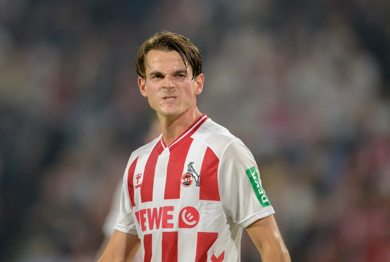 Piłkarz w stroju drużyny FC Köln z białymi i czerwonymi pasami stoi na boisku, wyrażając emocje, w tle widownia stadionu.