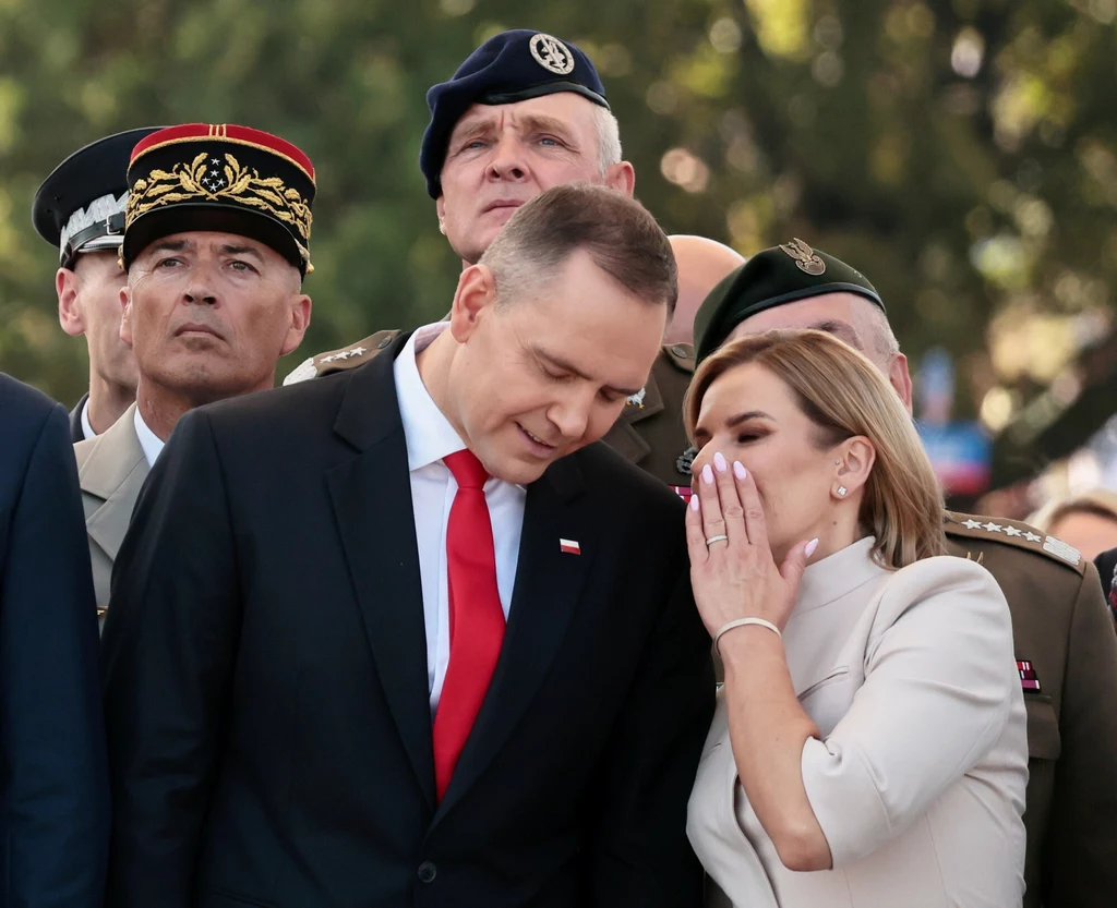 Marta Nawrocka podczas wystąpień publicznych prezentuje się w perfekcyjnych zestawach, a jej manicure akcentuje charakter całego wizerunku Grupa wysokich rangą wojskowych w mundurach oraz mężczyzna w garniturze ze znaczkiem flagi Polski i kobieta w jasnym stroju, która dyskretnie szepce coś mężczyźnie do ucha, w tle rozmyte drzewa.