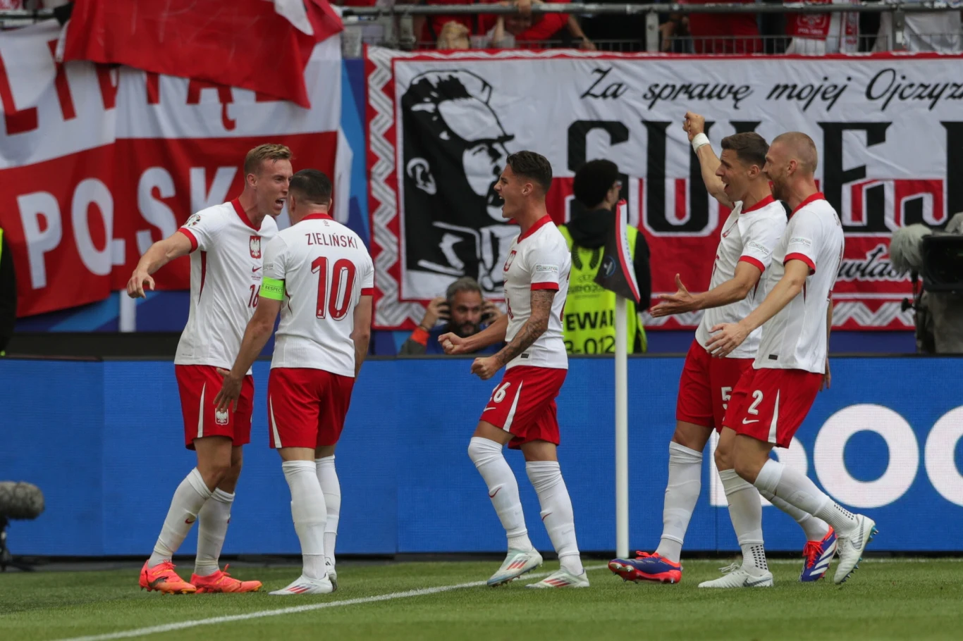 Reprezentacja Polski Grupa polskich piłkarzy świętuje zdobycie bramki na tle kibiców i flag narodowych, na boisku panuje atmosfera radości i jedności.