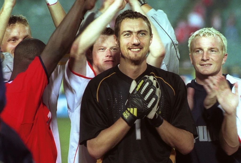 Polska - Norwegia. Na pierwszym planie Jerzy Dudek Grupa piłkarzy świętujących zwycięstwo na boisku, w centrum stoi bramkarz w czarnej koszulce z rękawicami, uśmiechnięty, wokół niego koledzy z drużyny wyrażają radość gestami i brawami.