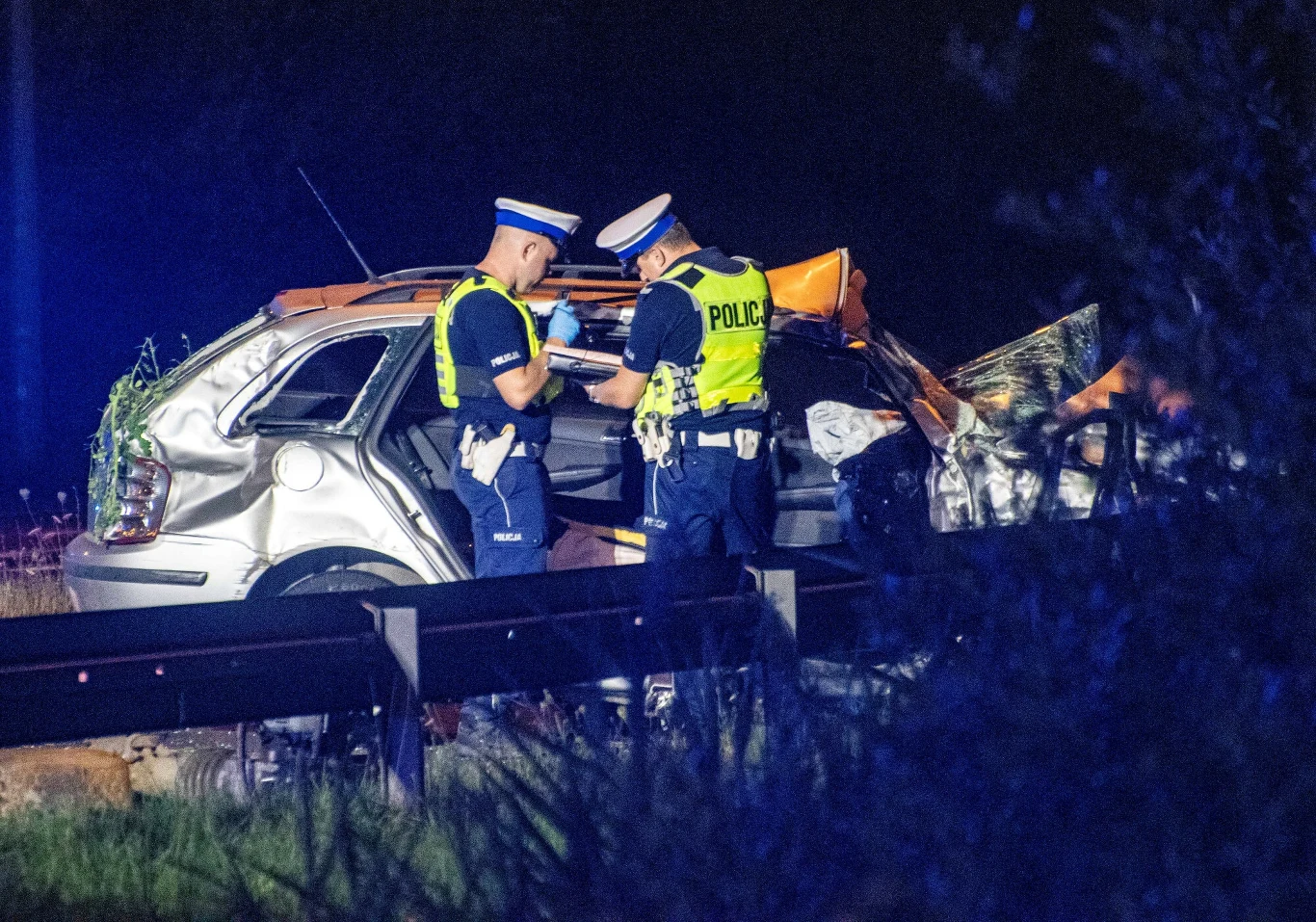 W wakacje w całej Polsce doszło do ponad 4,6 tys. wypadków drogowych Dwóch funkcjonariuszy policji w odblaskowych kamizelkach stoi obok mocno zniszczonego samochodu, nocna scena oświetlona światłami policyjnymi, po lewej stronie widoczny fragment barierki ochronnej.