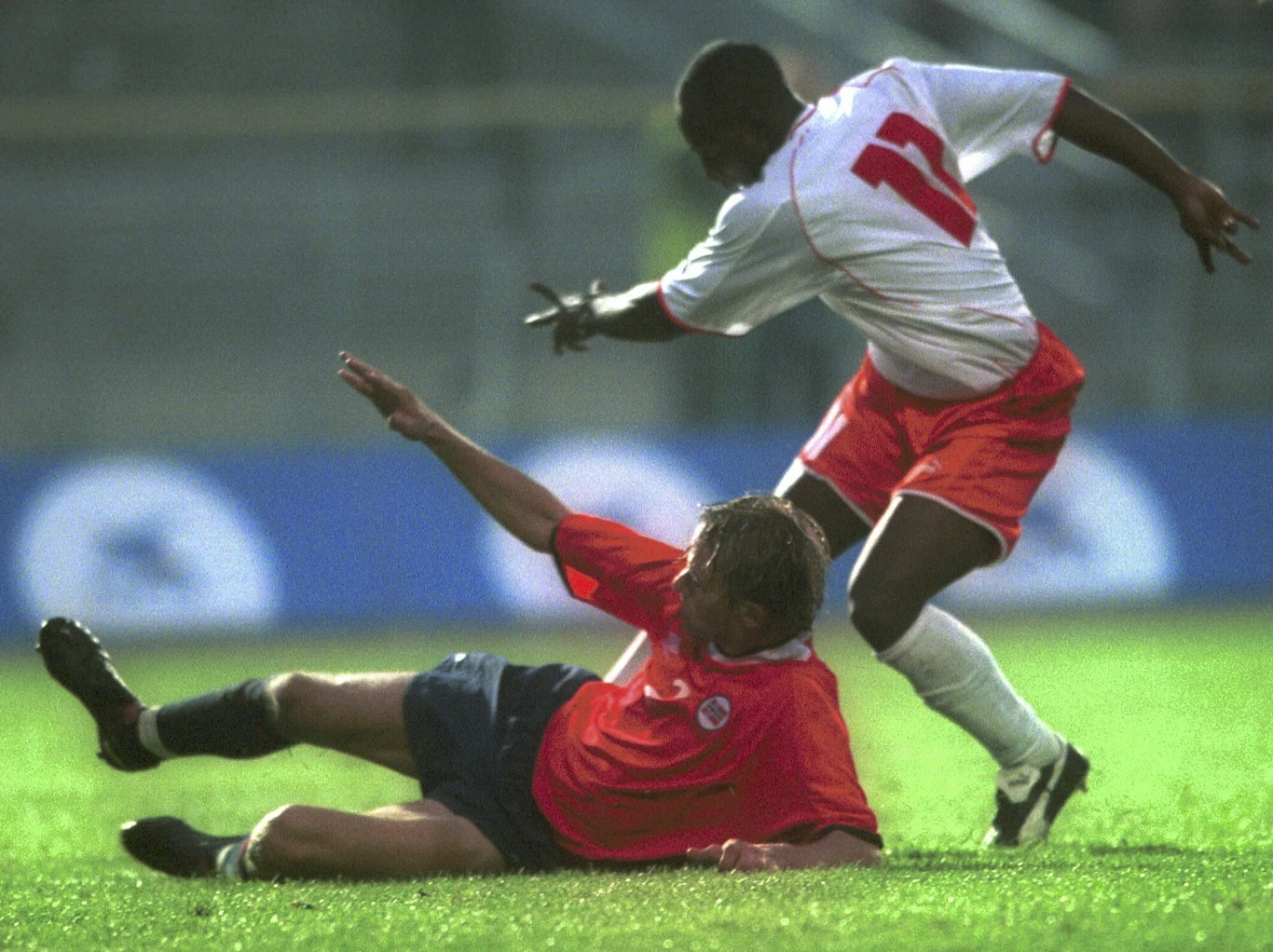 Dwóch piłkarzy w intensywnym starciu o piłkę na murawie, jeden z zawodników podczas wślizgu próbuje przejąć piłkę, drugi utrzymuje równowagę i dynamicznie reaguje na akcję przeciwnika