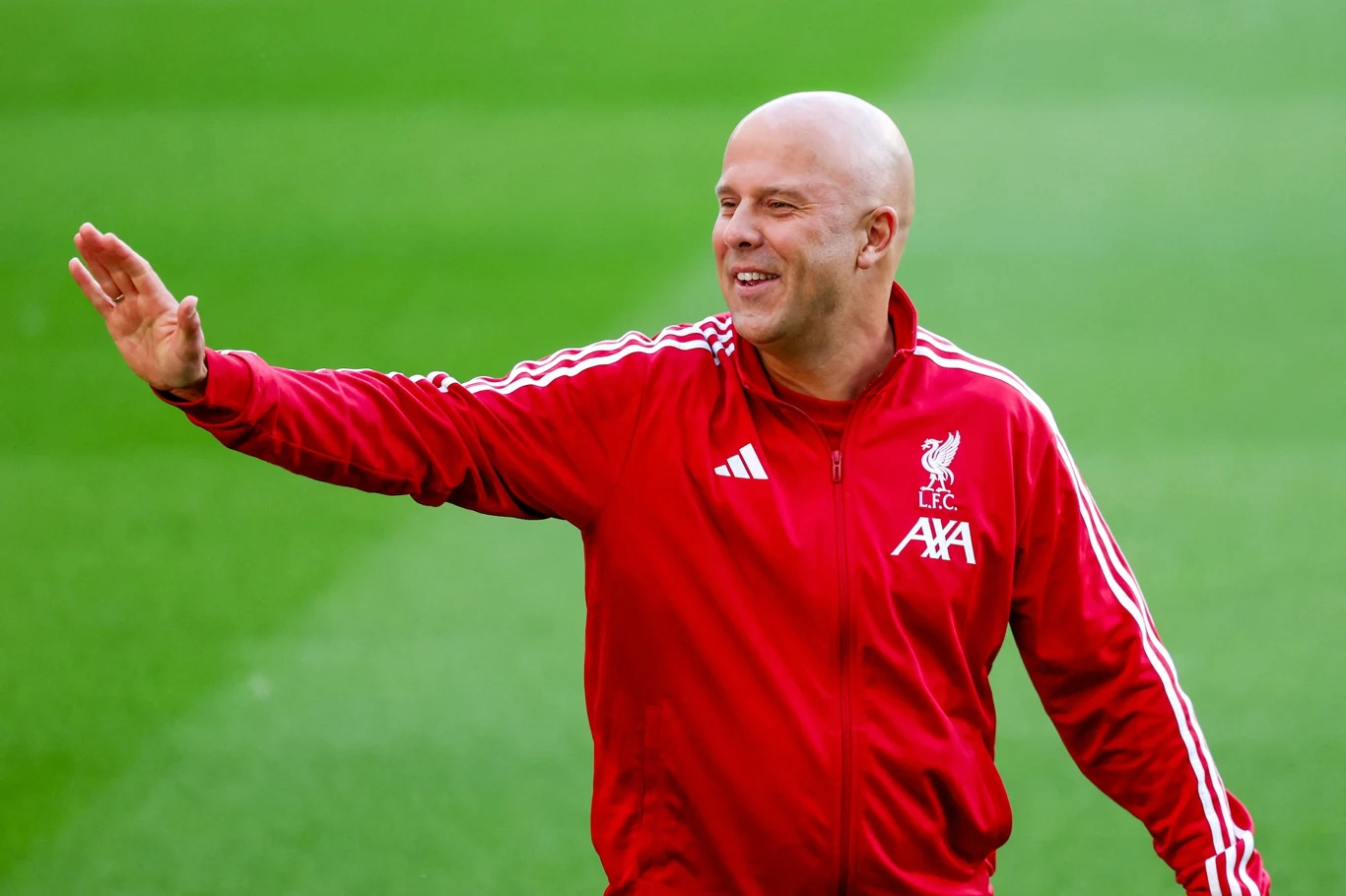 Arne Slot Mężczyzna w czerwonym dresie z logo Liverpool FC uśmiecha się i macha ręką na zielonym boisku sportowym.