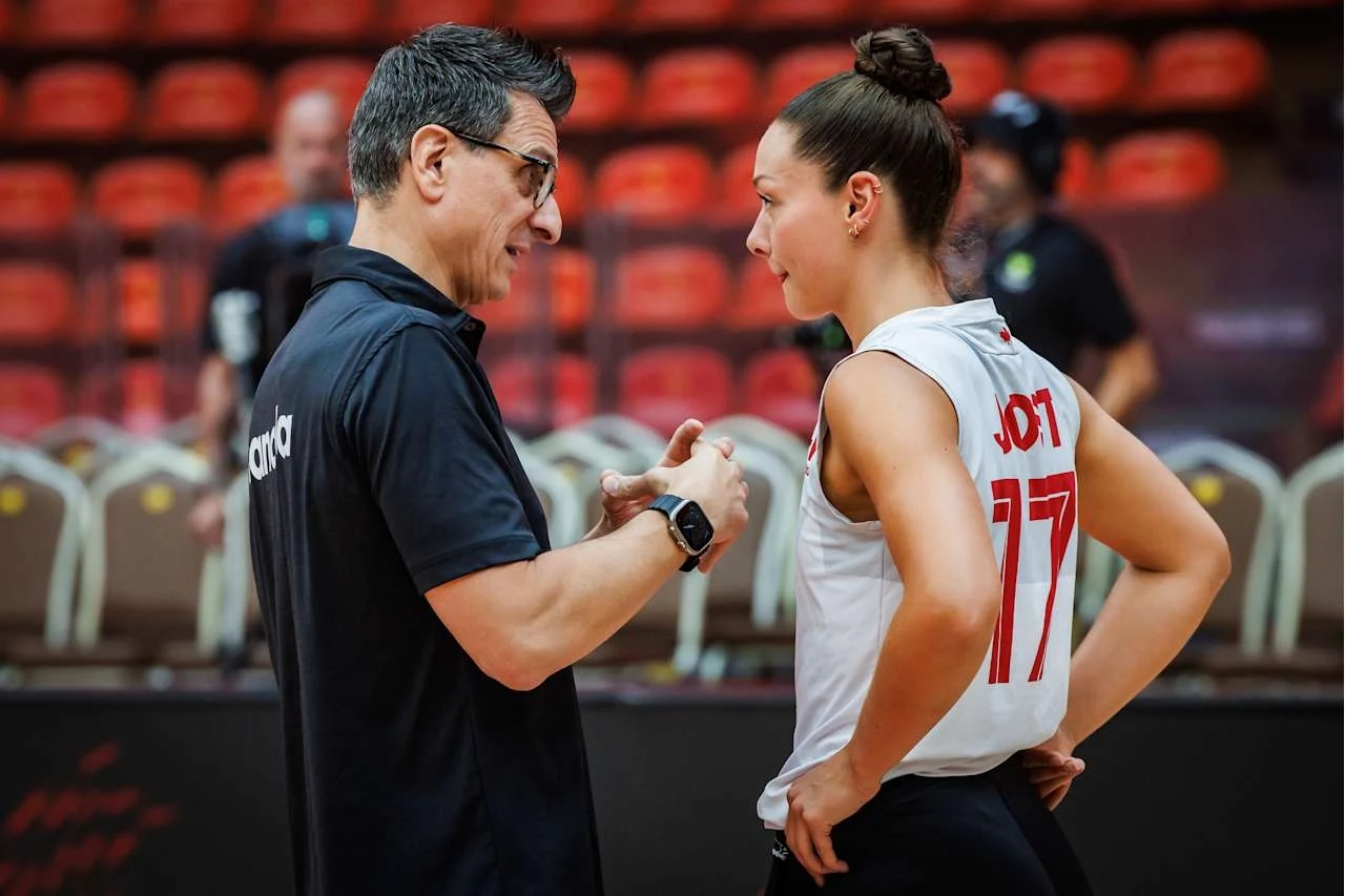 Trener rozmawia z siatkarką w sportowej hali, oboje skoncentrowani, wokół widoczne są puste krzesła i rozmyte sylwetki innych osób.