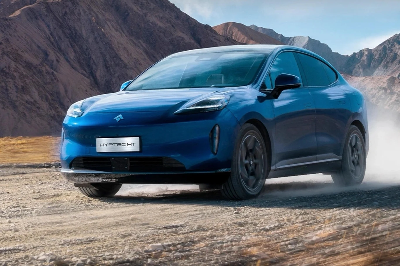 Niebieski samochód elektryczny typu SUV marki HYPTCE HT jedzie po szutrowej drodze w górskim, suchym krajobrazie, w tle widać szczyty gór i pył unoszący się spod kół.