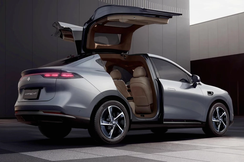 Wyróżnikiem modelu Hyptec HT będą unoszone do góry drzwi Srebrny samochód elektryczny z otwartymi tylnymi drzwiami i bagażnikiem, zaparkowany na nowoczesnym, miejskim placu; wnętrze pojazdu wykończone beżową skórzaną tapicerką.