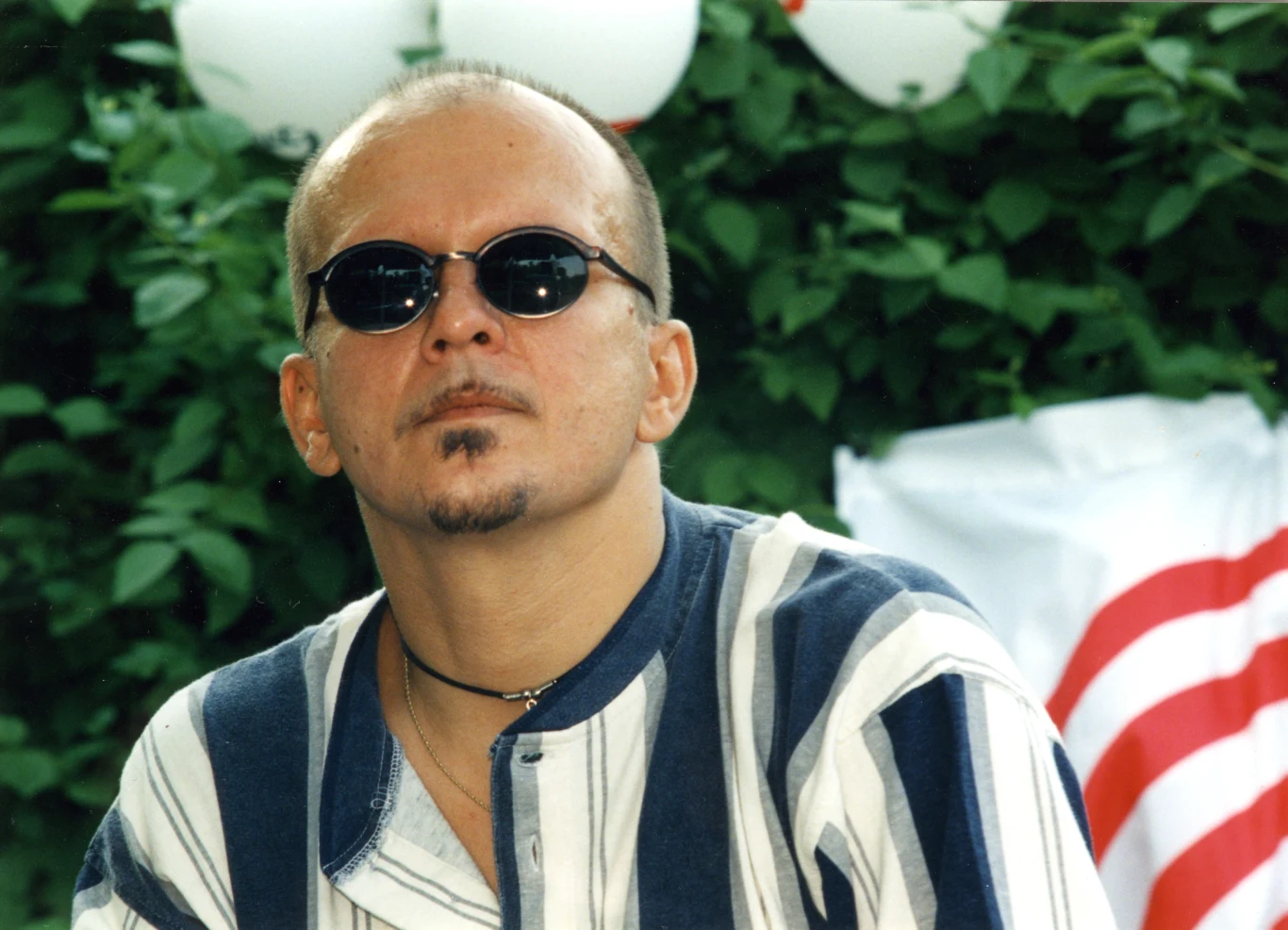 Ryszard Sygitowicz (na zdjęciu w 1996 r.) był muzykiem pierwszego składu grupy Perfect Mężczyzna w okrągłych okularach przeciwsłonecznych, ubrany w koszulę w biało-granatowe pasy, z krótkimi włosami i niewielką bródką, siedzi na tle zielonych liści oraz balonów i fragmentu flagi z czerwonymi paskami.