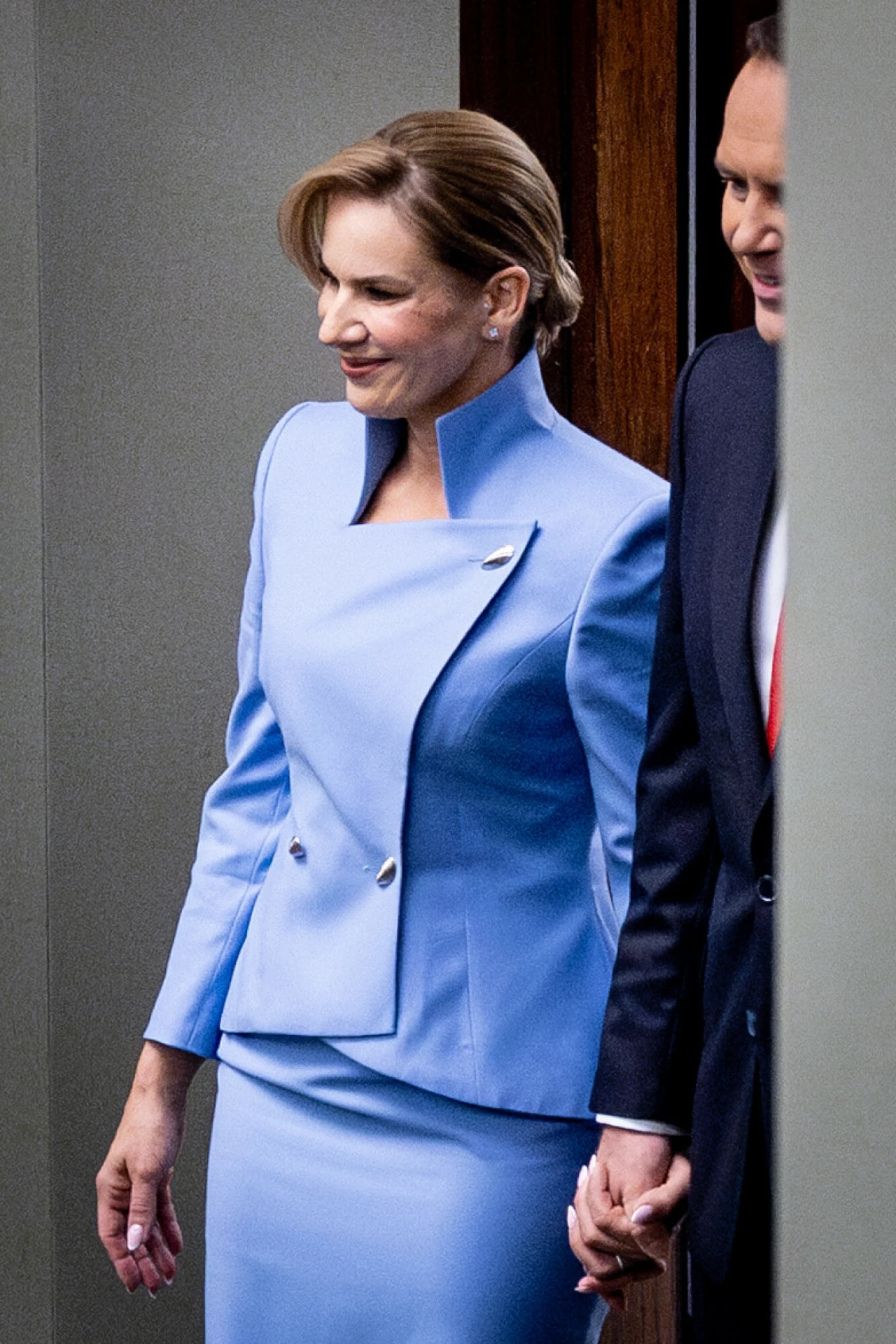 Ta błękitna stylizacja zapadła w pamięć, choć nie była krzykliwa Elegancko ubrana kobieta w jasnoniebieskim kostiumie trzyma za rękę mężczyznę w ciemnym garniturze, oboje wychodzą z pomieszczenia, subtelny uśmiech na twarzy kobiety, formalna atmosfera.