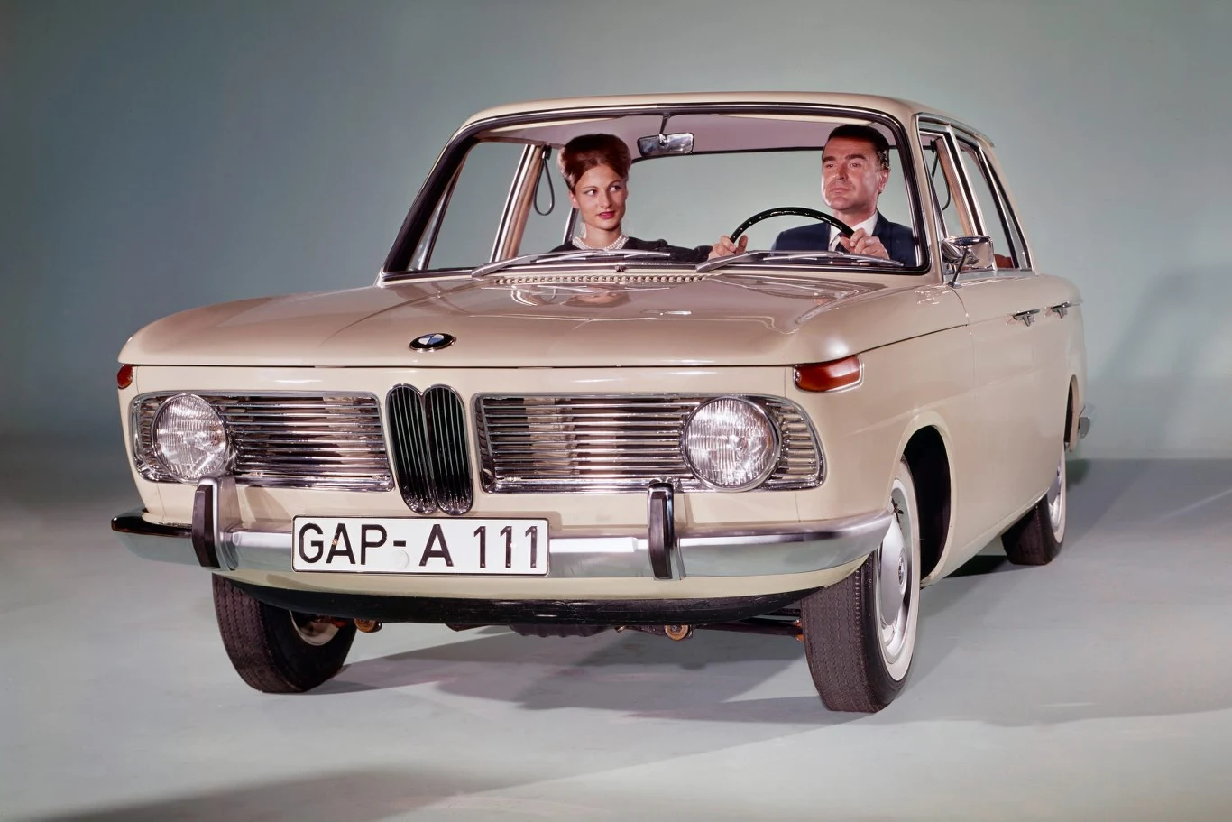 Klasyczny samochód BMW z lat 60., w jasnym kolorze, na pierwszym planie; za kierownicą elegancko ubrany mężczyzna, obok niego kobieta; tło jednolite, studyjne.