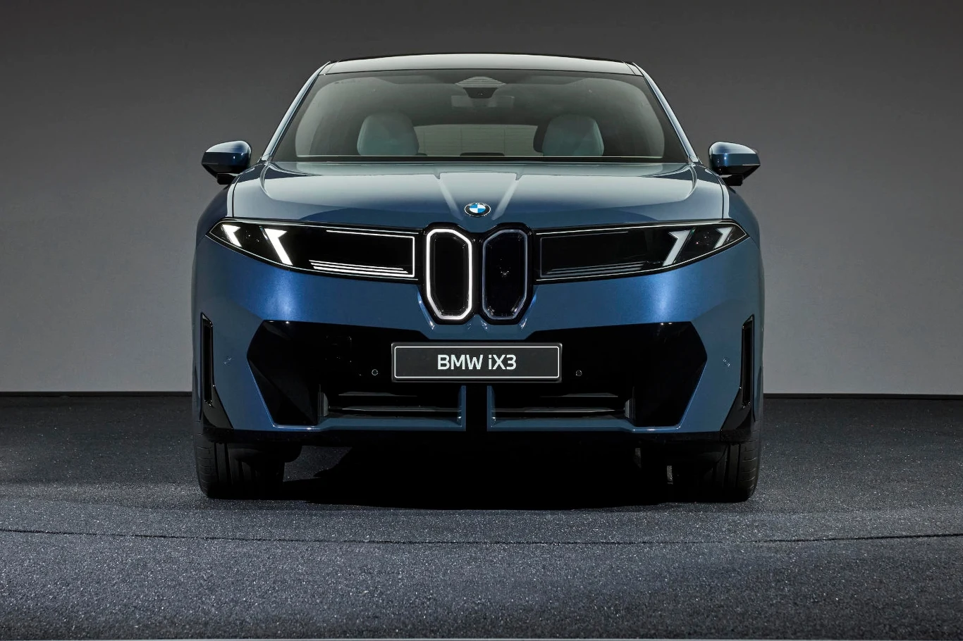 Nowoczesny elektryczny samochód BMW iX3 w kolorze niebieskim sfotografowany od frontu na ciemnym tle, z wyraźnie widocznym charakterystycznym grillem i reflektorami.