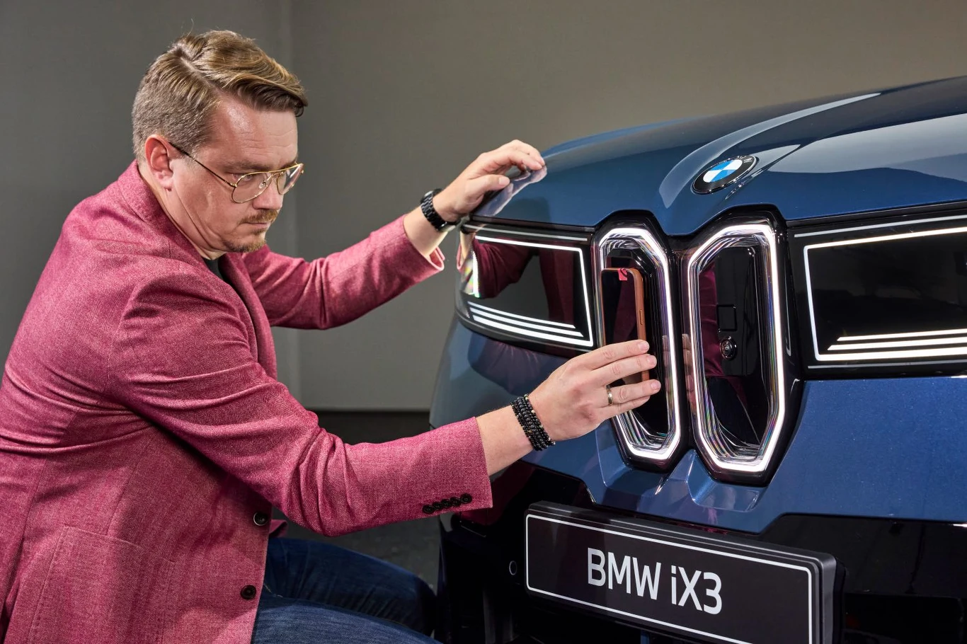 Mężczyzna w okularach i różowej marynarce dotyka przedniego grilla nowoczesnego, niebieskiego samochodu BMW iX3, koncentrując się na detalach emblematu i kształtu reflektorów.