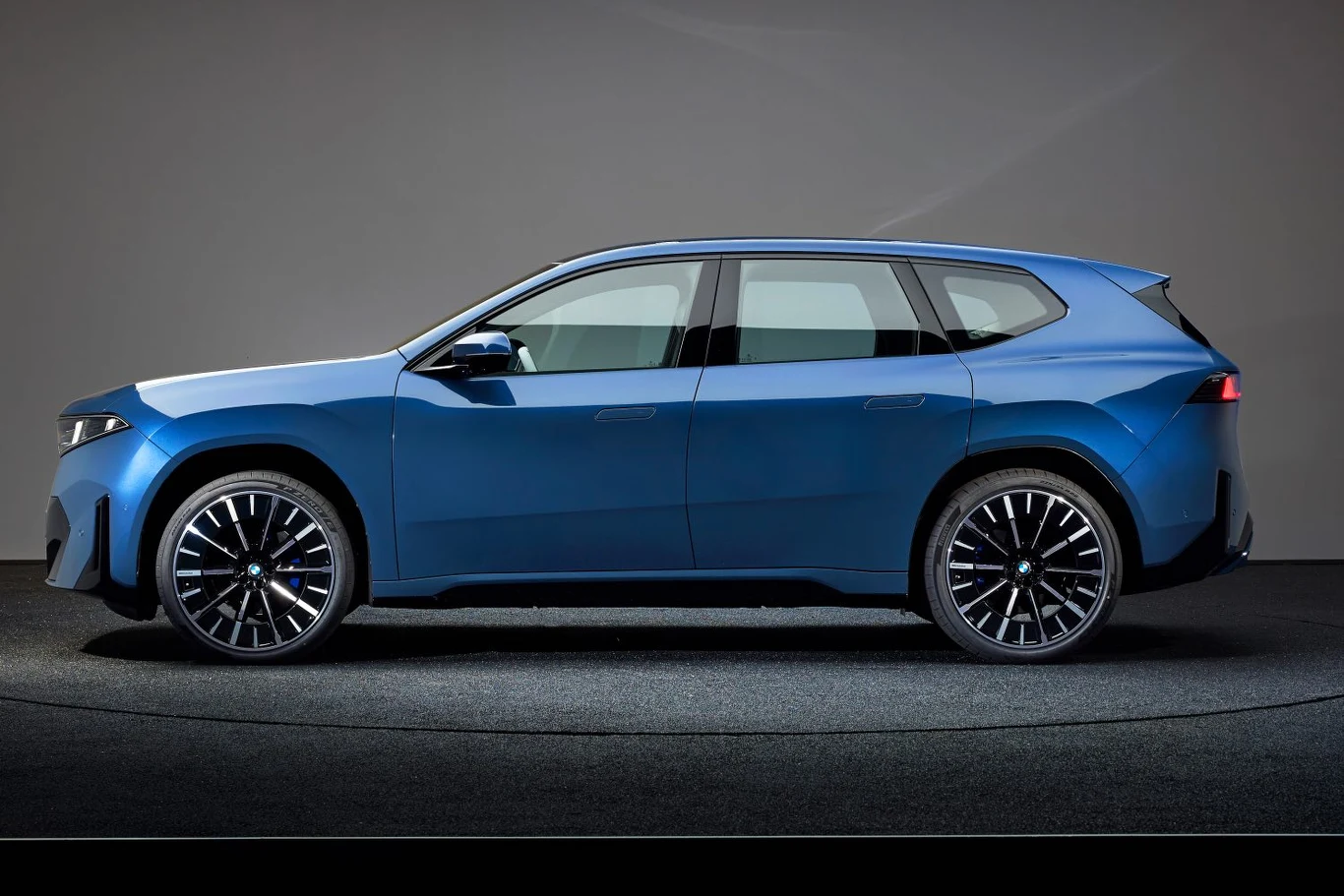 Nowoczesny, niebieski samochód typu SUV widoczny z profilu na tle neutralnej, jednolitej ściany, auto stoi na ciemnym dywanie oświetlonym punktowo, ma wyraźnie zarysowane linie nadwozia i ciemne, wieloramienne felgi.