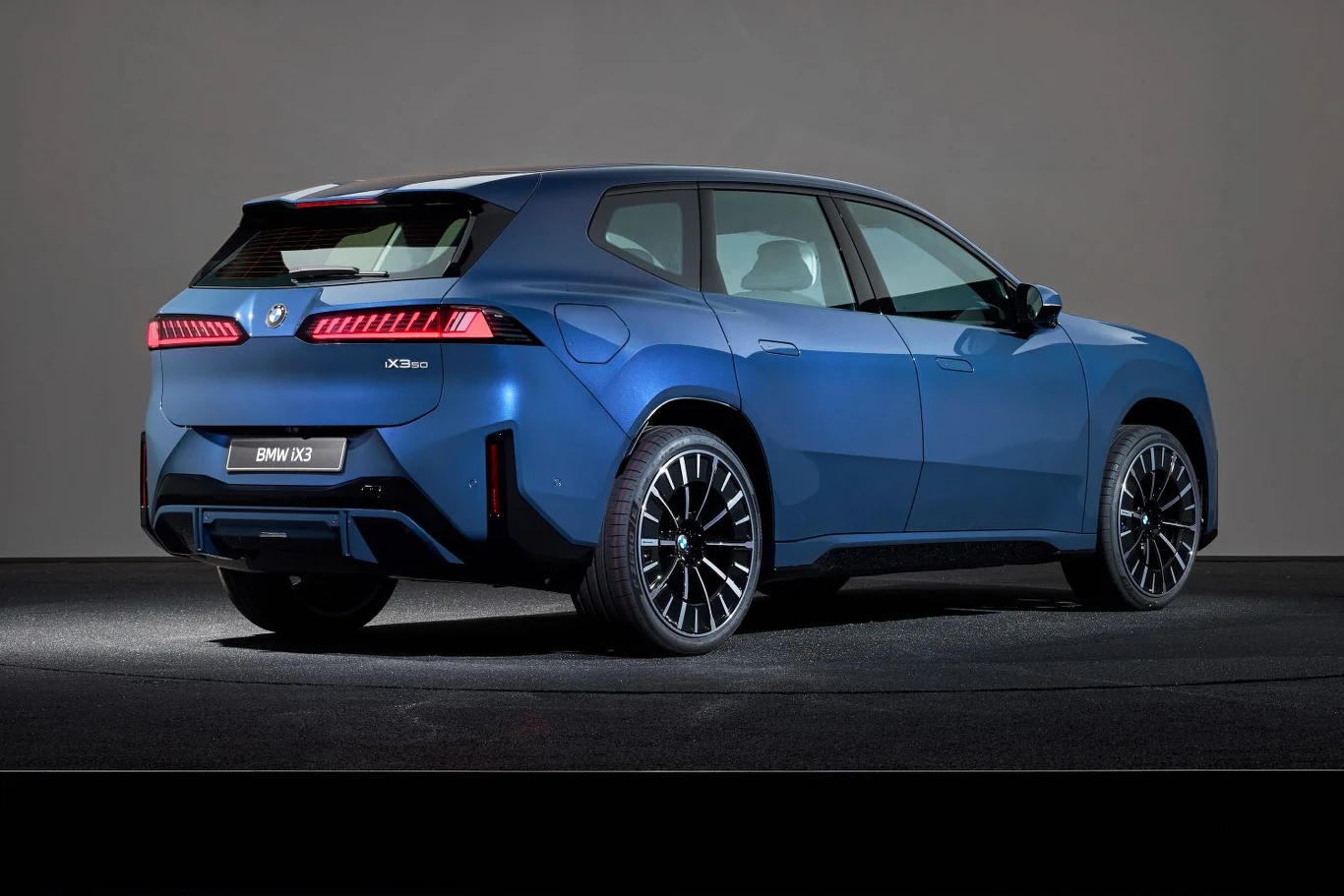 Nowoczesny, niebieski SUV BMW X3 widziany z tyłu, wyeksponowane dynamiczne linie nadwozia, jasne tylne światła LED, wyraźne logo marki i napisy modelu, auto stoi na czarnej nawierzchni w neutralnym otoczeniu.