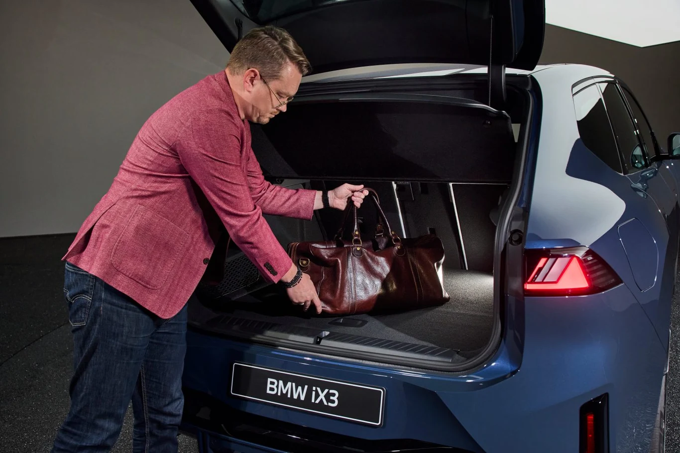 Mężczyzna w różowej marynarce wkłada skórzaną torbę podróżną do bagażnika niebieskiego samochodu BMW iX3, bagażnik jest otwarty, wnętrze dobrze oświetlone.