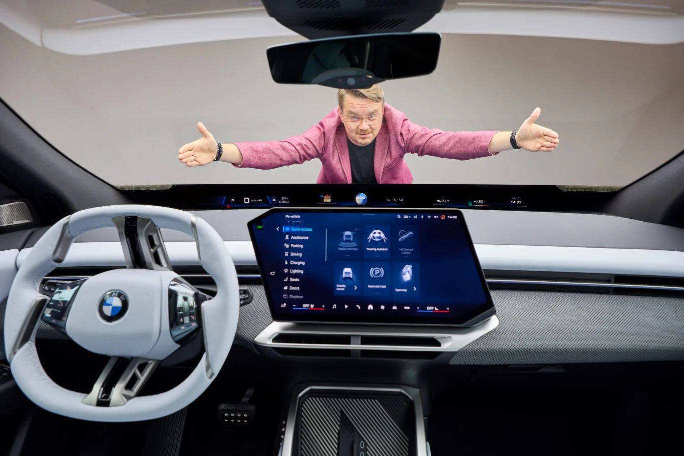 Widok nowoczesnego wnętrza samochodu BMW z cyfrowym wyświetlaczem na desce rozdzielczej i dużym ekranem dotykowym, za szybą stoi mężczyzna w różowej marynarce z szeroko rozpostartymi rękami, jakby tłumaczył lub gestykulował.