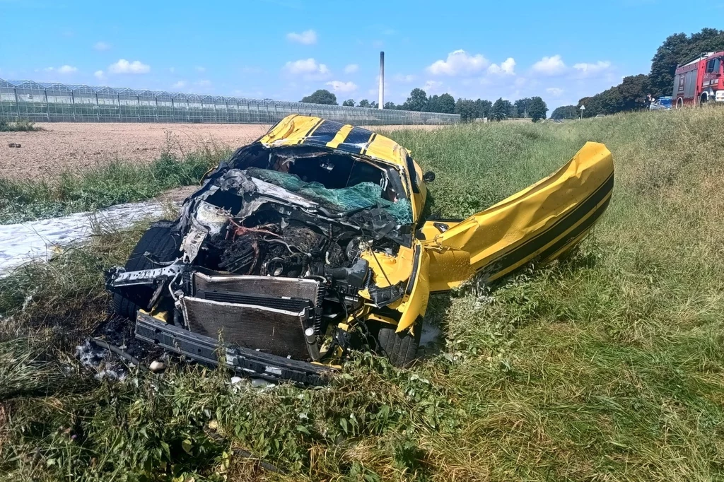 Rozbity żółty samochód leży w rowie przy drodze, zniszczony przód oraz otwarte, zdeformowane drzwi, w tle widoczny fragment pola, ogrodzenie i wóz strażacki.