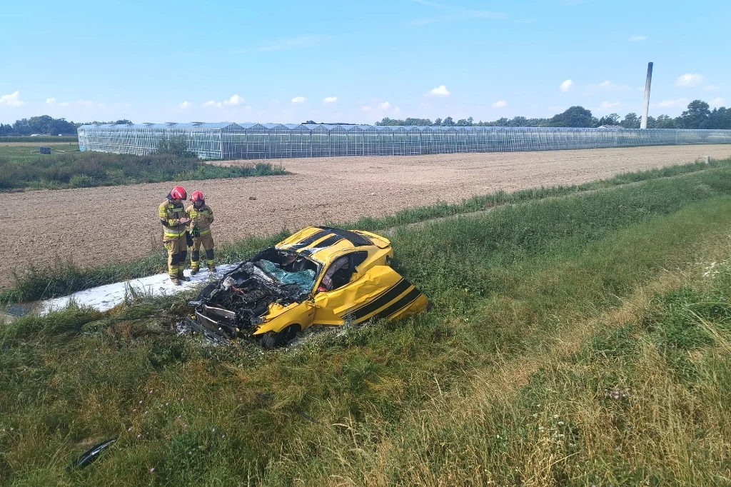Rozbity żółty samochód osobowy leży w rowie przy polnej drodze, obok stoją dwaj strażacy w pełnym umundurowaniu, tło stanowią pola uprawne oraz szklarnie.