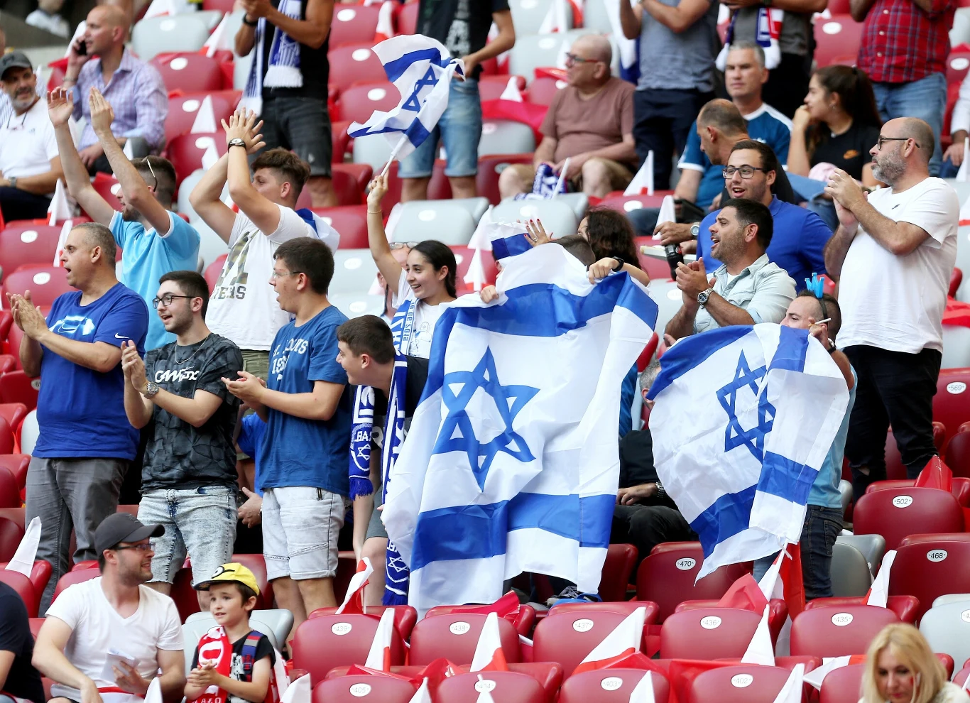 Tłum kibiców na stadionie trzymających flagi Izraela, część osób klaszcze i wyraża entuzjazm, na trybunach dominuje atmosfera wsparcia dla drużyny sportowej.