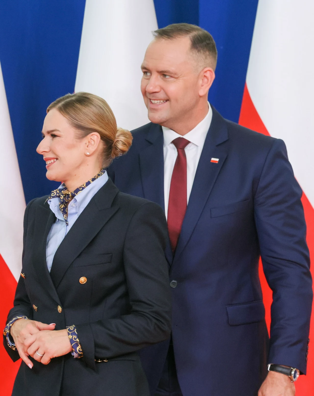 Marta Nawrocka i Karol Nawrocki. Para prezydencka pojawiła się na rozpoczęciu roku szkolnego w Wolanowie Para elegancko ubranych osób stojąca na tle polskich flag, mężczyzna w garniturze z biało-czerwoną plakietką w klapie i kobieta w ciemnym żakiecie, oboje uśmiechnięci, w oficjalnym otoczeniu.