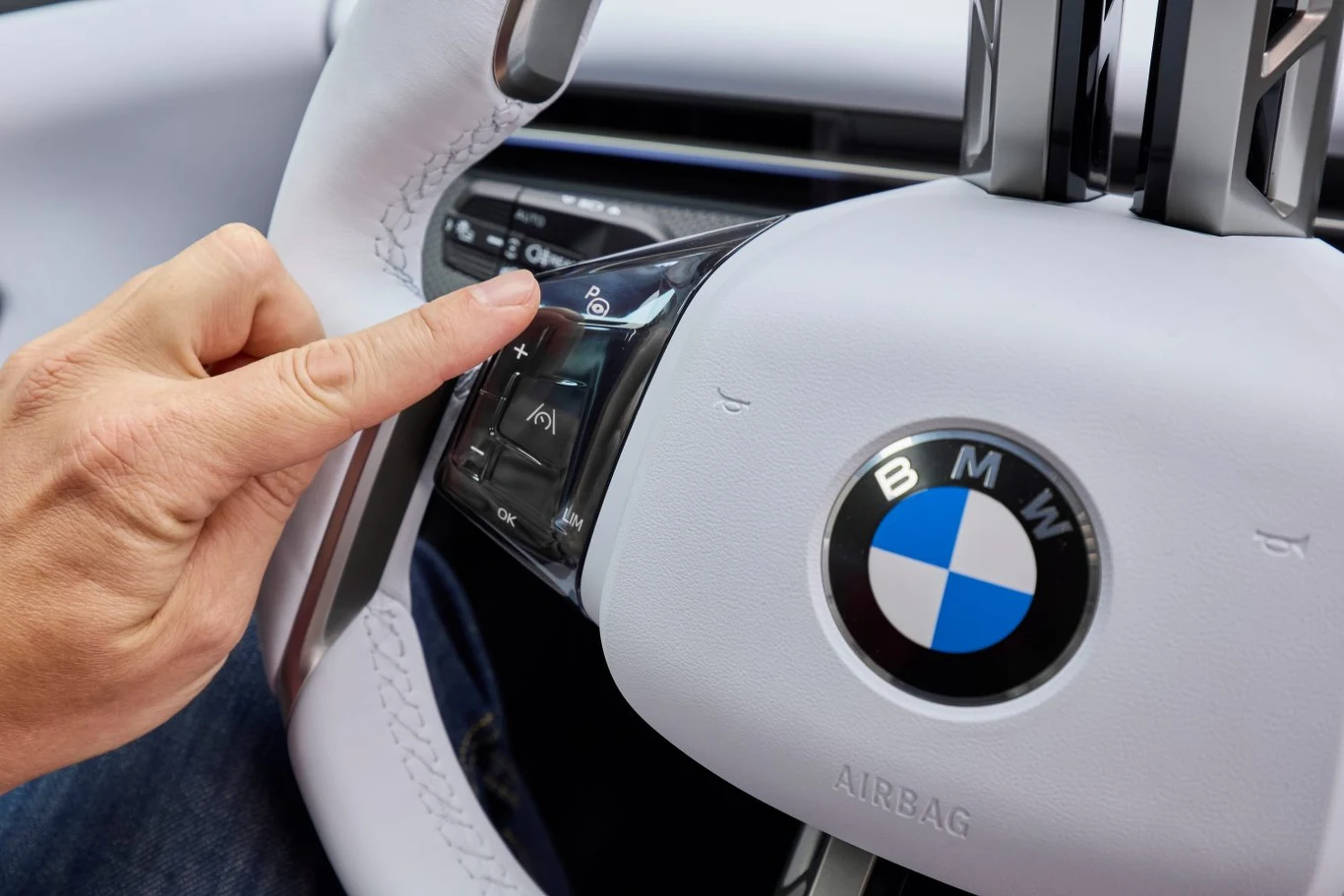Ręka osoby naciskająca przycisk na kierownicy samochodu marki BMW z widocznym znakiem firmowym i panelem do sterowania funkcjami pojazdu.