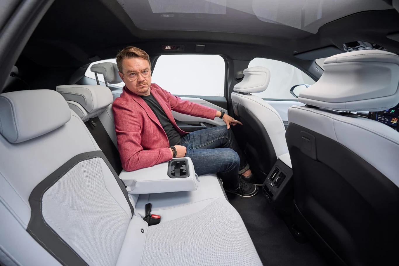 Mężczyzna w okularach i czerwonej marynarce siedzi na tylnym siedzeniu nowoczesnego samochodu z jasnym, skórzanym wnętrzem, opierając rękę na podłokietniku i spoglądając w stronę aparatu.