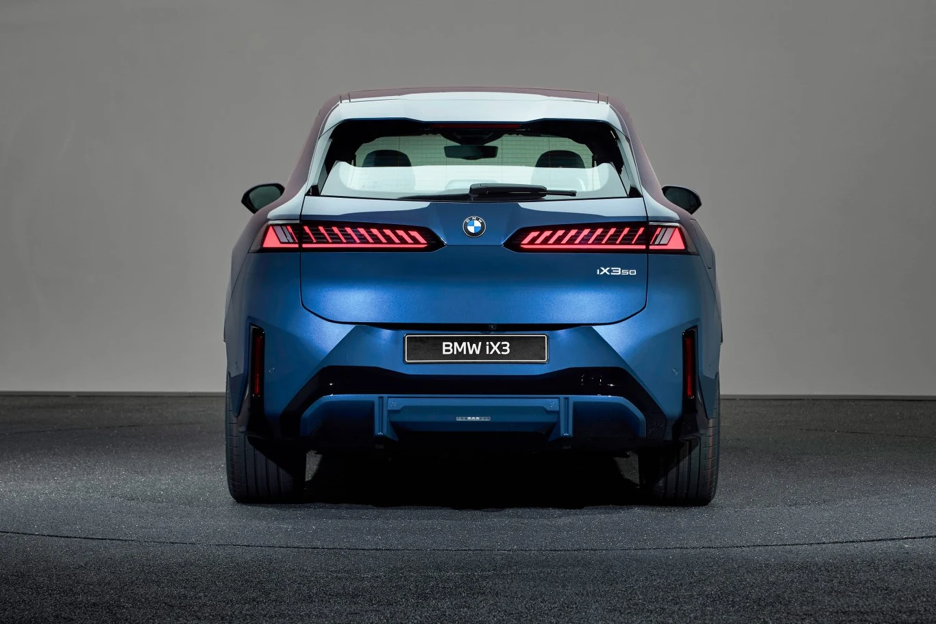 Nowoczesny elektryczny SUV BMW IX3 w niebieskim kolorze widoczny od tyłu, z podświetlonymi, wąskimi lampami oraz charakterystycznym logo BMW, ustawiony na neutralnym tle.