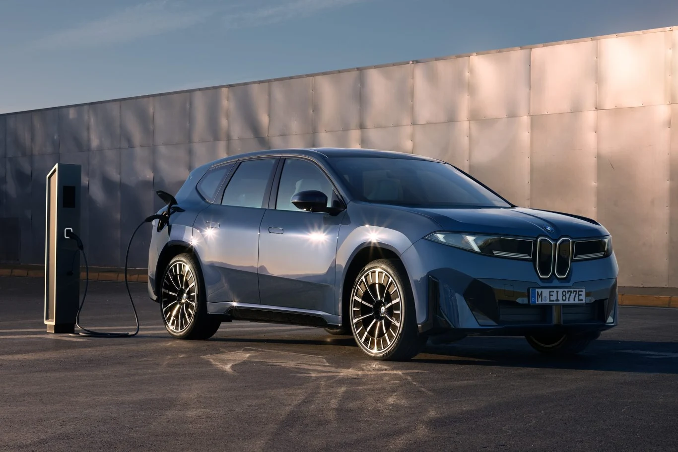 Nowoczesny, niebieski samochód elektryczny marki BMW stoi przy stacji ładowania na otwartym, nasłonecznionym parkingu, kabel ładowarki podłączony do pojazdu.