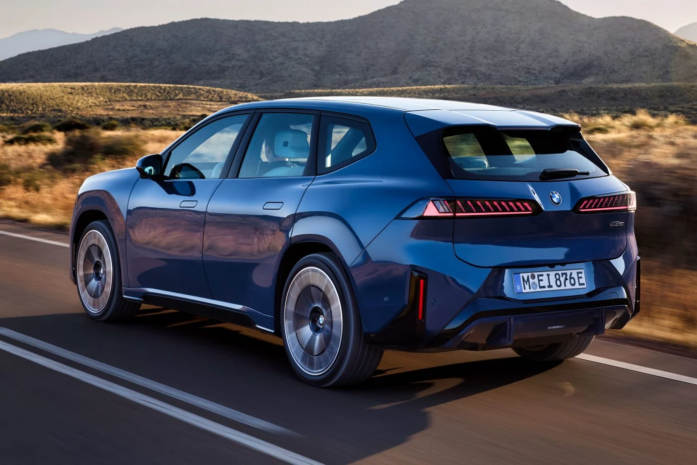 Nowoczesny, niebieski samochód marki BMW jedzie po asfaltowej drodze przez suchy, górzysty krajobraz, przy słonecznej pogodzie.