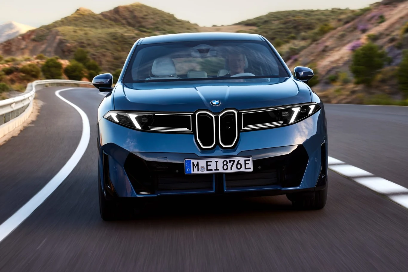 Nowoczesny, niebieski samochód marki BMW jedzie dynamicznie po krętej, górskiej drodze, otoczonej zielenią i skałami, a zdjęcie wykonane jest z przodu auta, podkreślając jego agresywny przód i reflektory.