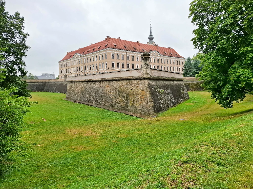 Zamek Lubomirskich w Rzeszowie – twierdza z poczatku XVI w. Historyczna budowla z czerwonym dachem na murach obronnych otoczona szerokim, zielonym trawnikiem oraz drzewami po bokach, w tle pojedyncze drzewa oraz fragmenty nowoczesnych budynków.