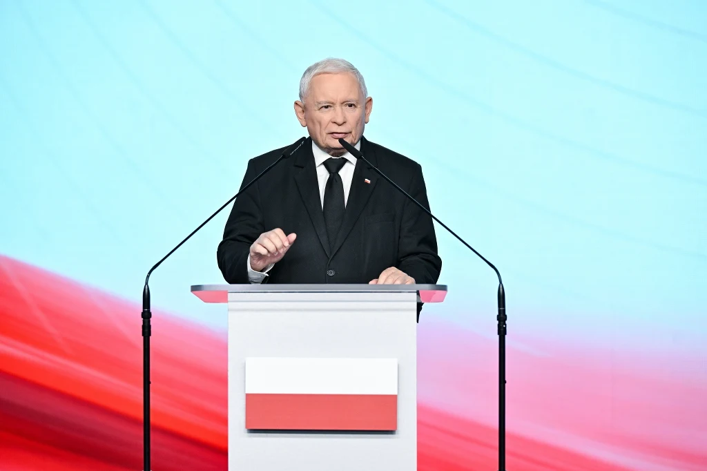 Jarosław Kaczyński apeluje do rządzących ws. mrożenia cen prądu Starszy mężczyzna w eleganckim, czarnym garniturze przemawia stojąc za przezroczystą mównicą z polską flagą, w tle znajduje się jasnoniebieskie i czerwone tło przypominające kolory flagi Polski.