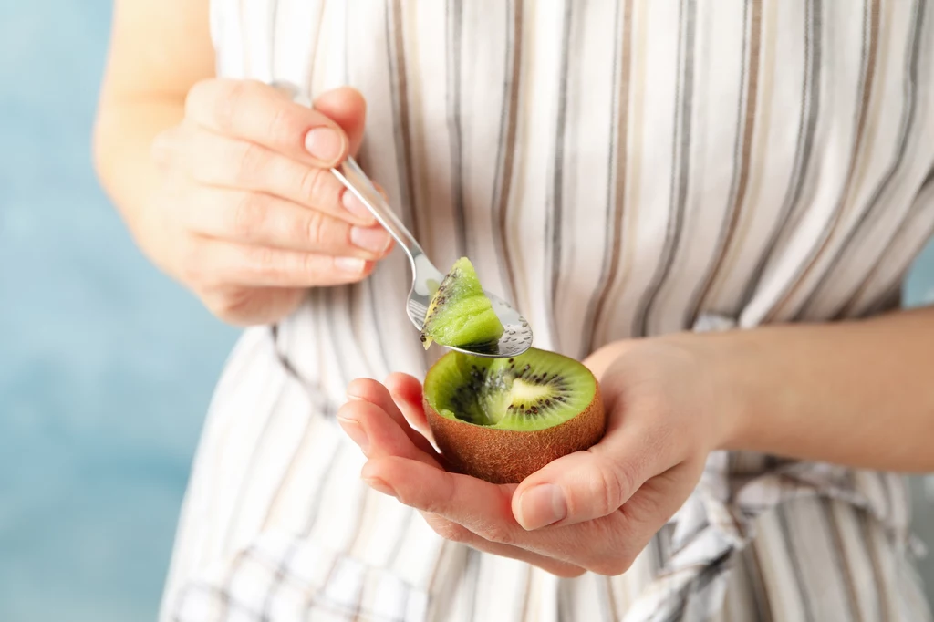 Jak jeść kiwi ze skórką? Osoba wybiera miąższ z połówki kiwi łyżeczką, trzymając owoc w dłoni i mając na sobie pasiasty fartuch.
