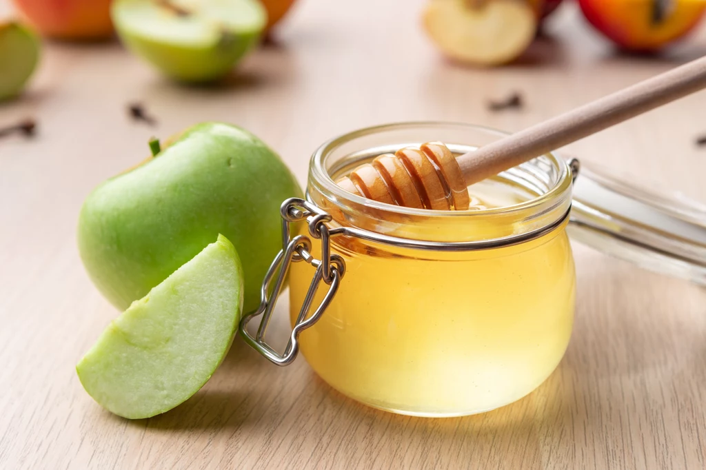 Maseczka z jabłka i miodu. Naturalny sposób na promienną cerę Słoik miodu z drewnianym nabierakiem, obok świeże zielone jabłko przekrojone na kawałki, w tle rozmyte jabłka i goździki.