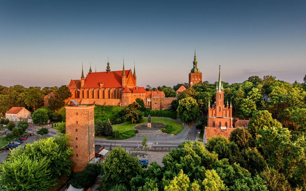 Gdzie na jednodniowy trip? Poznaj najciekawsze polskie miasta dla seniorów Ceglany zespół katedralny otoczony zielonymi drzewami, dominująca gotycka katedra z licznymi wieżyczkami, wieża oraz mniejsze obiekty sakralne w historycznym centrum miasta, spokojna przestrzeń miejska oświetlona popołudniowym światłem.