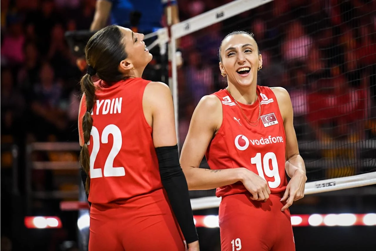 Dwie siatkarki w czerwonych strojach sportowych stojące blisko siatki, uśmiechnięte oraz wyraźnie zadowolone, tło rozmyte z obecnością widzów na trybunach.