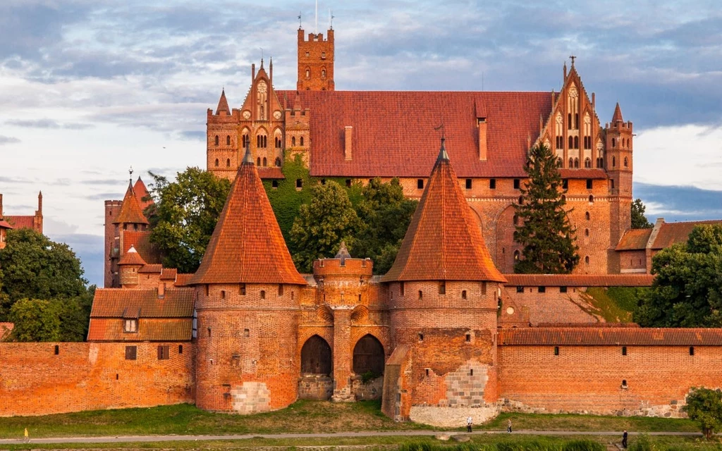 Zamek w Malborku Ceglany zamek otoczony masywnymi murami i basztami, z charakterystycznymi czerwonymi dachami oraz centralną bramą wjazdową, usytuowany pośród zieleni, z piękną architekturą gotycką.