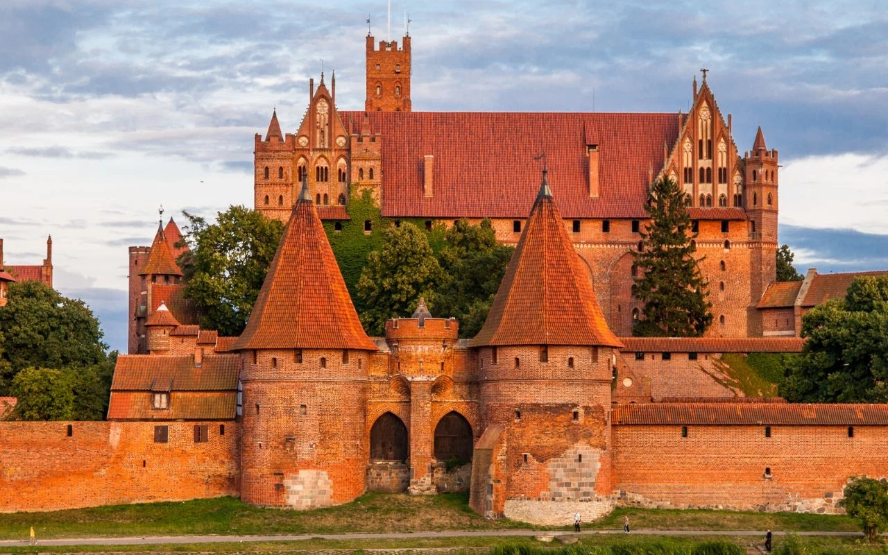 Zamek w Malborku Ceglany zamek otoczony masywnymi murami i basztami, z charakterystycznymi czerwonymi dachami oraz centralną bramą wjazdową, usytuowany pośród zieleni, z piękną architekturą gotycką.